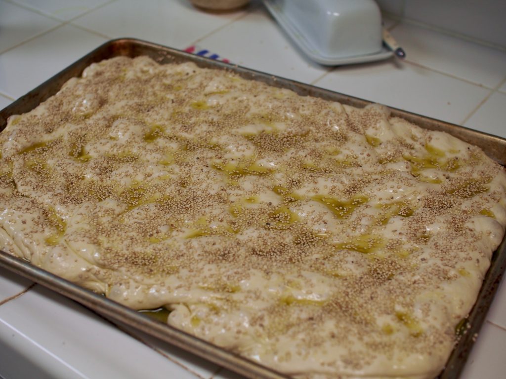 The uncooked focaccia is in a pan, glistening with oil and sesame seeds.
