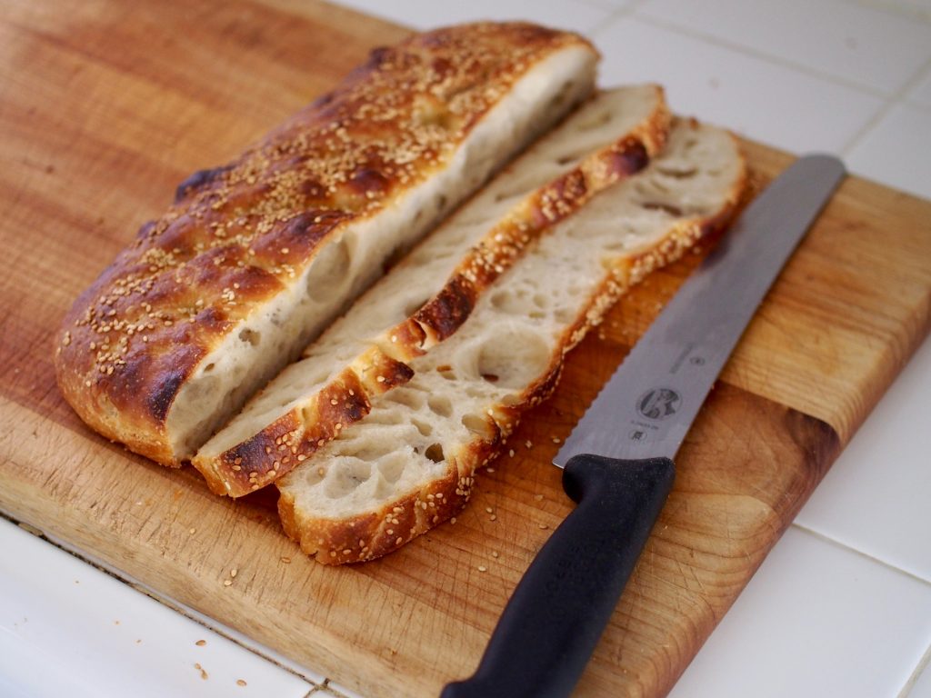 Two long, cross-wise slices of the focaccia rest on a board next to the heel of the bread. A black-handled bread knife sits to the right.
